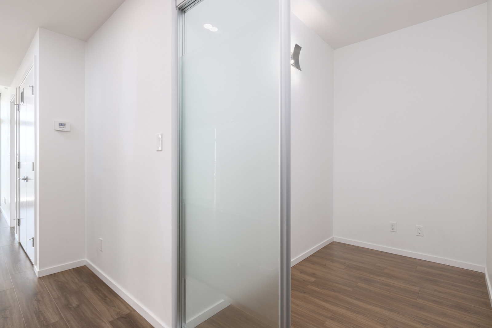 A small, empty room with white walls, wood flooring, and a frosted glass sliding door. Electrical outlets and a wall sconce are visible.