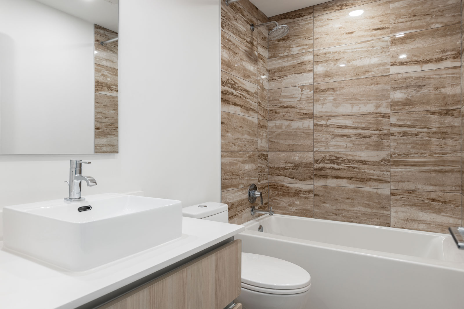 Modern bathroom with a white sink, countertop, toilet, and bathtub; wall behind the tub features brown marble tiles.