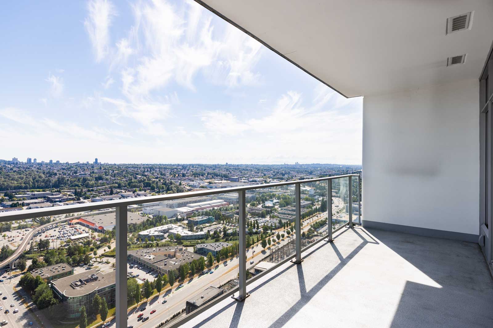High-rise balcony with glass railing overlooks a cityscape, including buildings, roads, and greenery under a bright sky with scattered clouds.