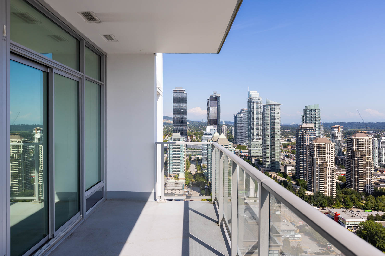 High-rise apartment balcony with glass railing, overlooking a cityscape of tall buildings under a clear blue sky.
