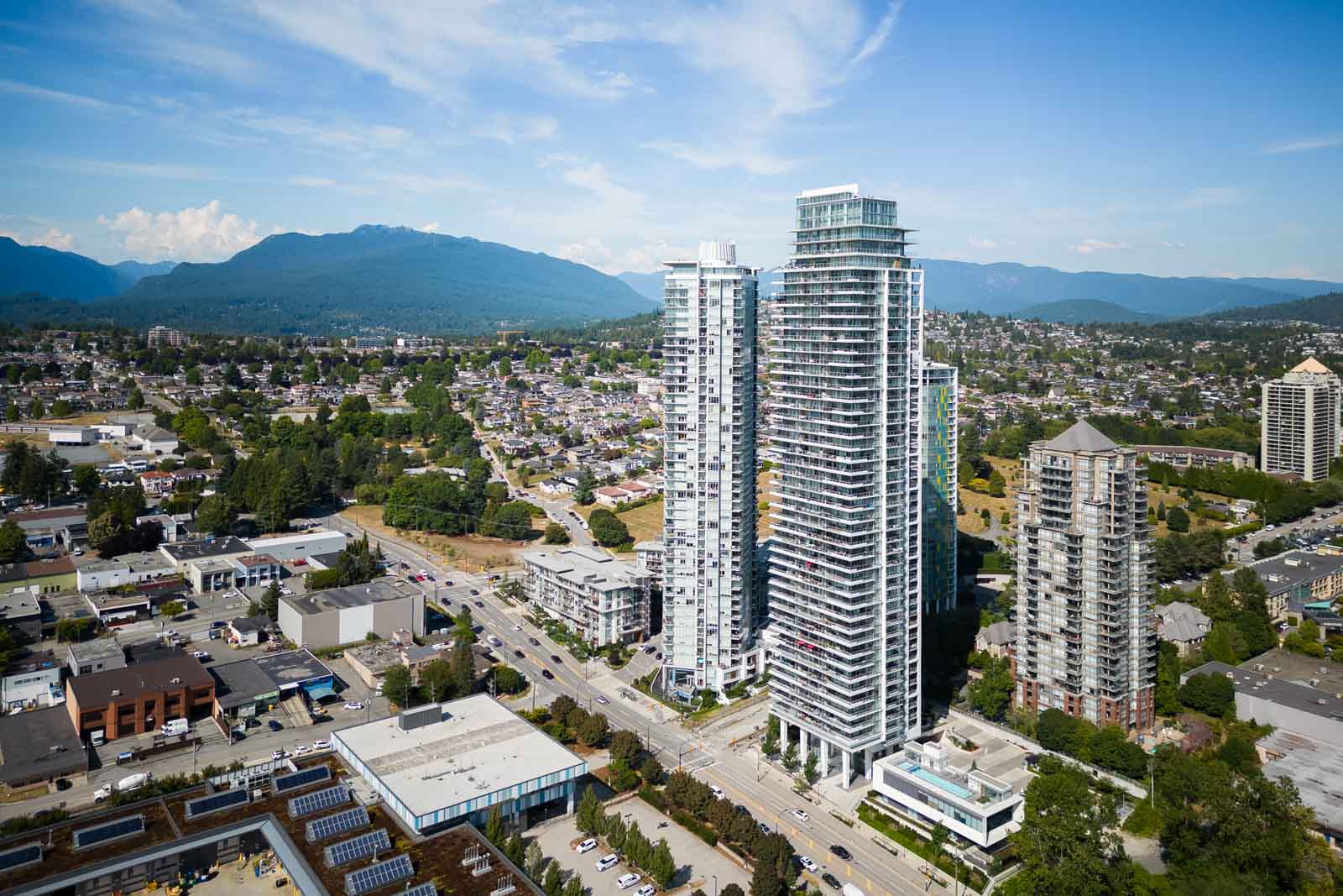 Aerial view of a cityscape with several tall modern apartment buildings, smaller commercial and residential structures, and mountains in the background under a partly cloudy sky.