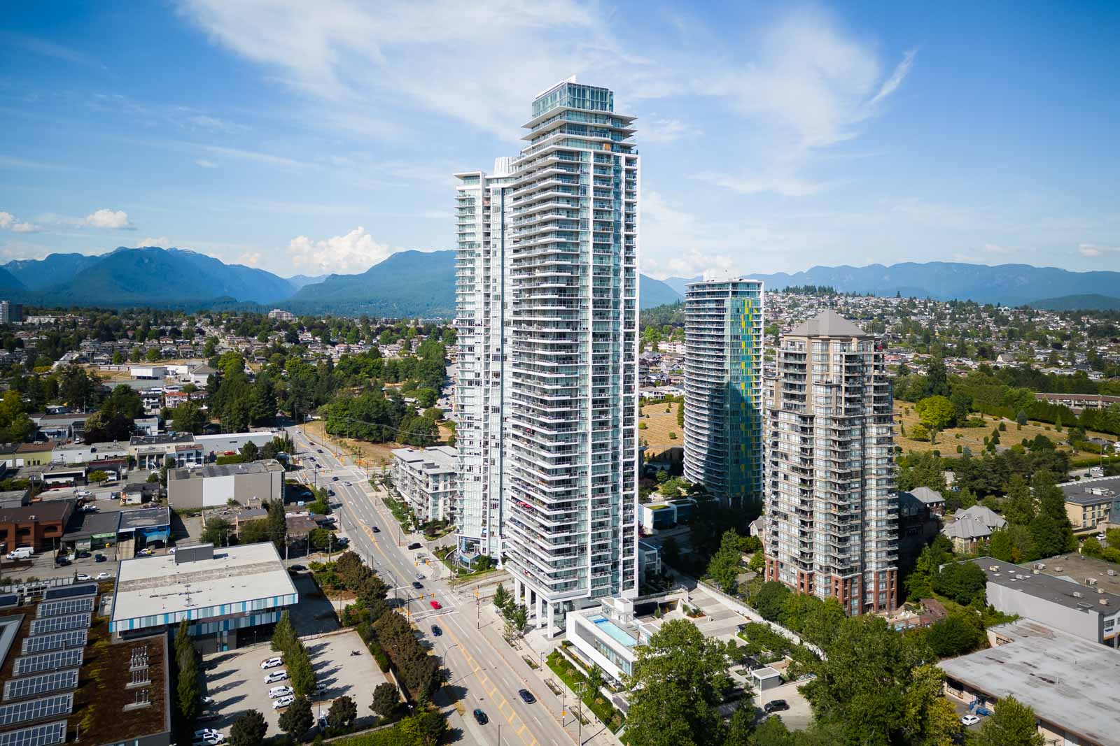 Aerial view of tall residential and commercial buildings along a busy road with mountains and blue sky in the background.