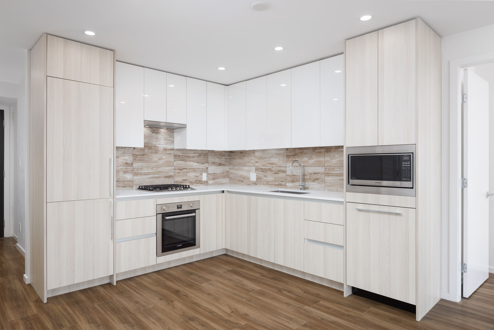 Modern kitchen with light wood cabinets, white countertops, built-in oven, microwave, gas stove, and neutral backsplash. Room has wood flooring and recessed ceiling lights.