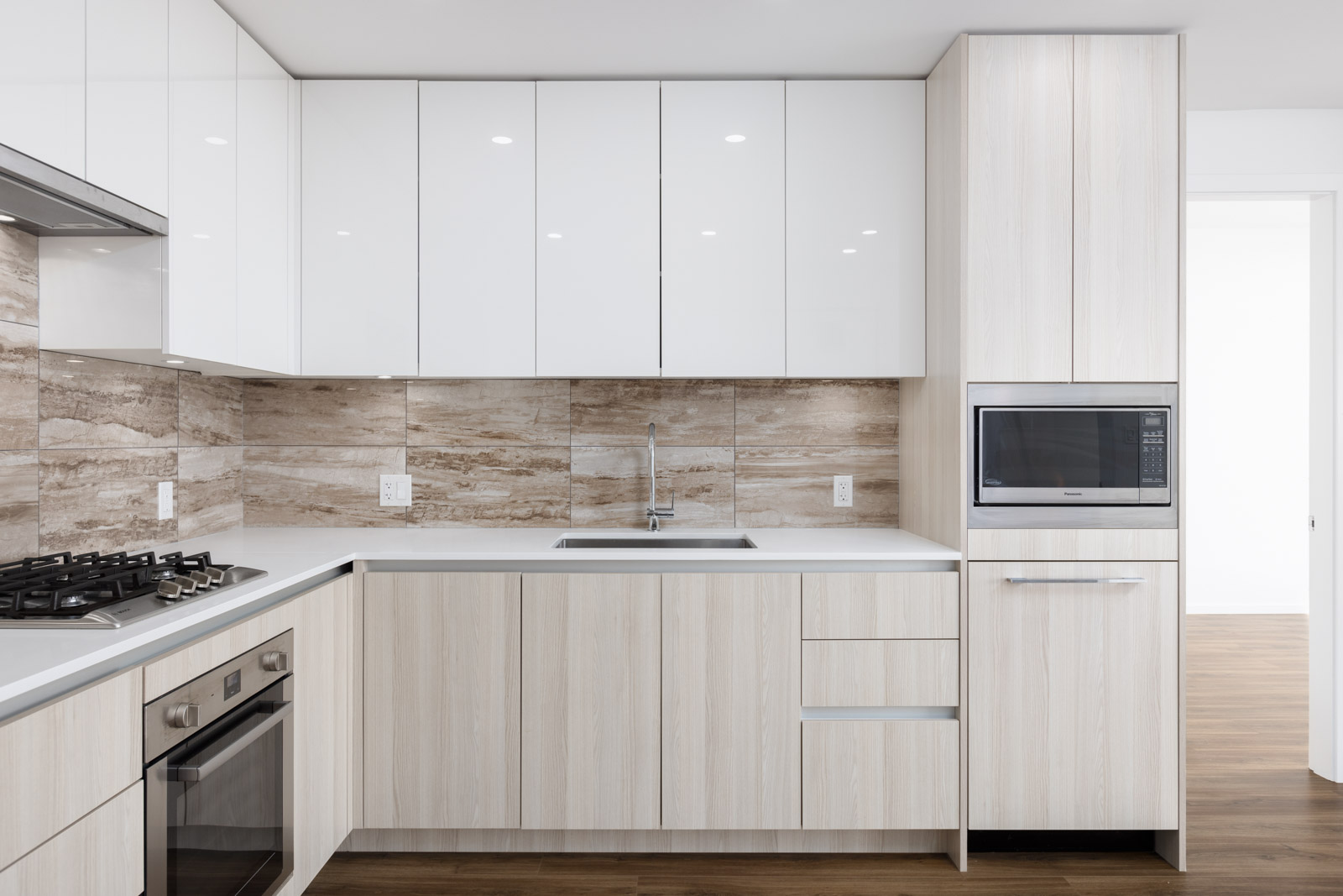 Modern kitchen with light wood cabinets, white countertops, built-in oven, gas stove, microwave, and a stainless steel sink against a beige tiled backsplash.
