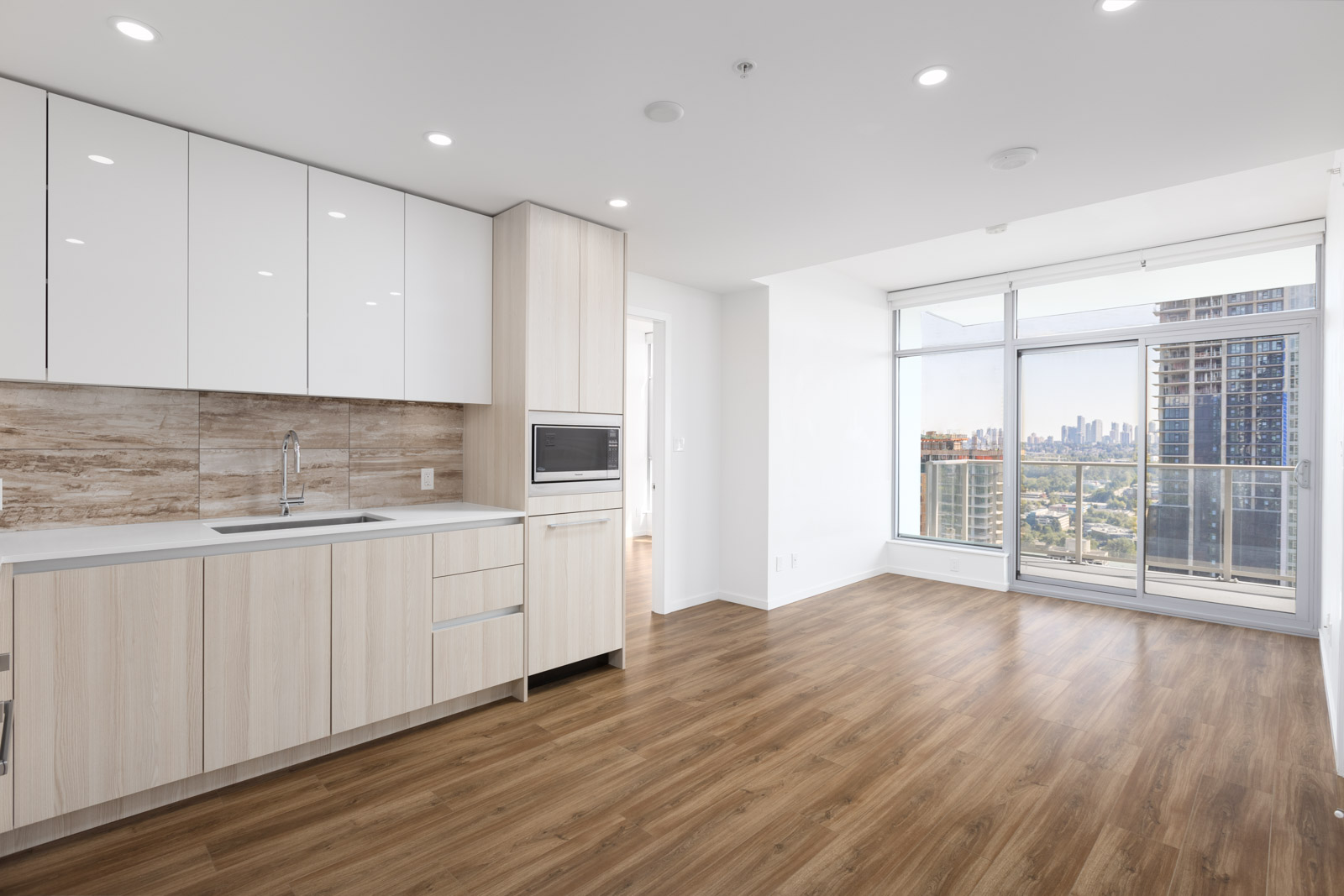 Modern apartment interior with light wood flooring, built-in kitchen cabinets, and large floor-to-ceiling windows offering a city view and access to a balcony.