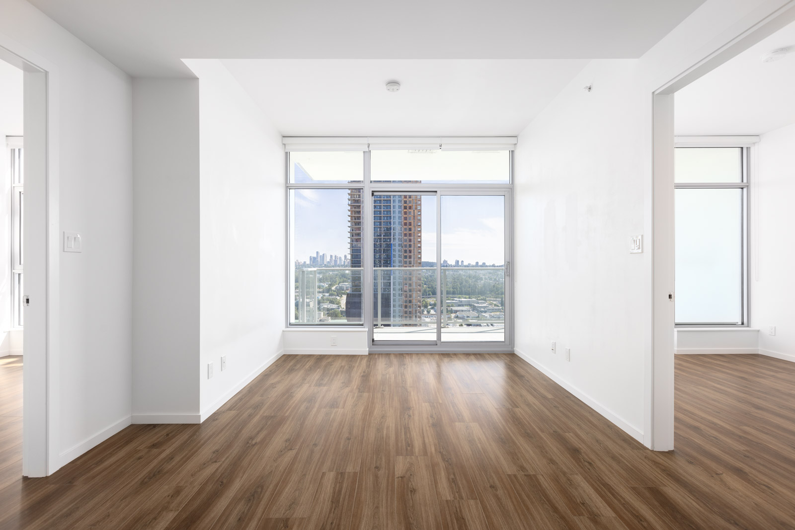 Empty, bright apartment room with white walls, wood flooring, large window, and glass balcony doors overlooking a cityscape; two open doorways on each side.