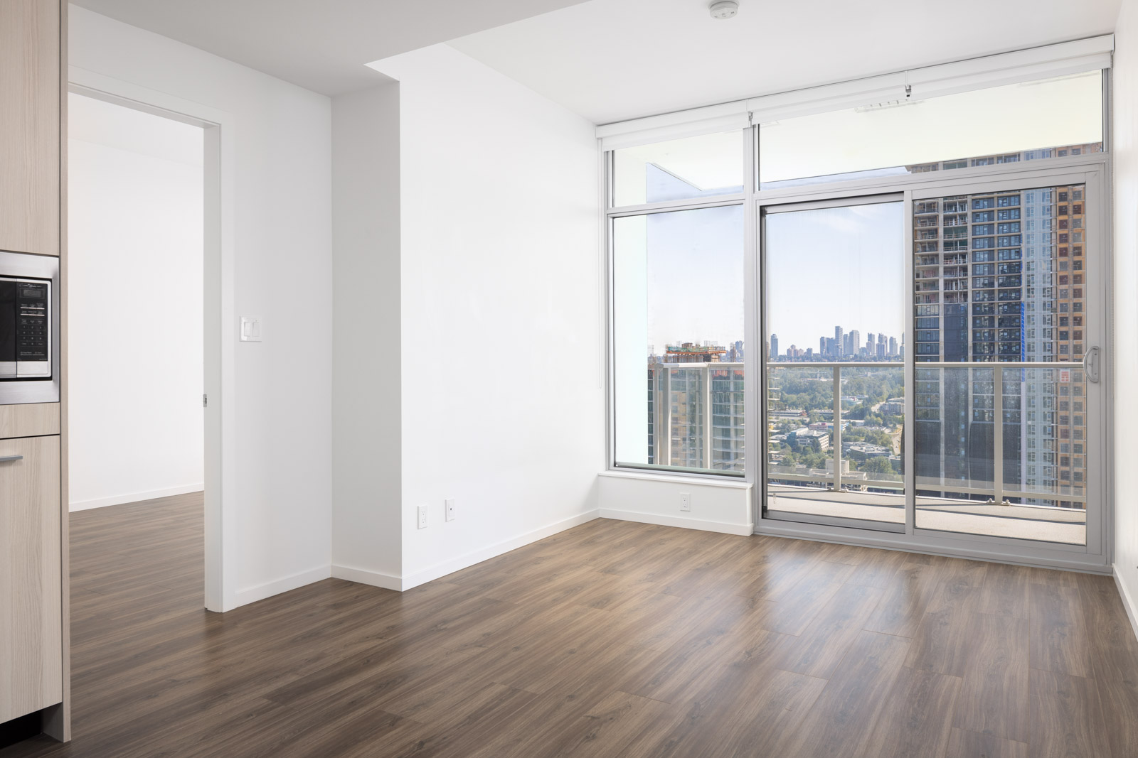 Bright empty apartment room with wood flooring, white walls, large windows, a glass door leading to a balcony, and a city skyline view in the background.