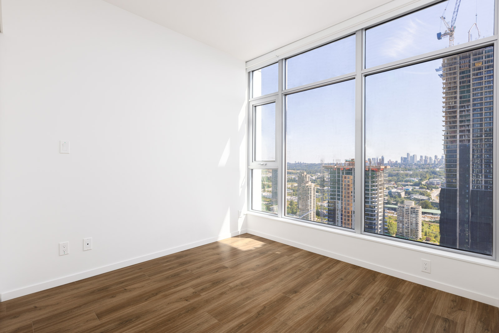 Empty room with white walls, large floor-to-ceiling windows, and wood flooring overlooking a cityscape with high-rise buildings and a distant skyline.