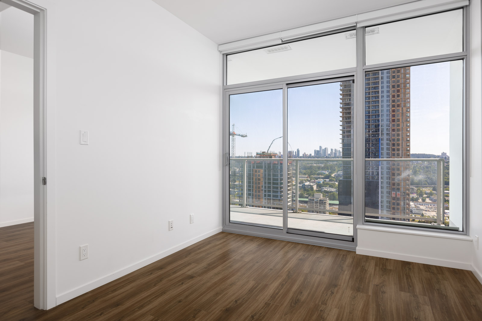 Unfurnished apartment room with white walls, wood flooring, and large glass doors leading to a balcony with a city view and tall buildings outside.