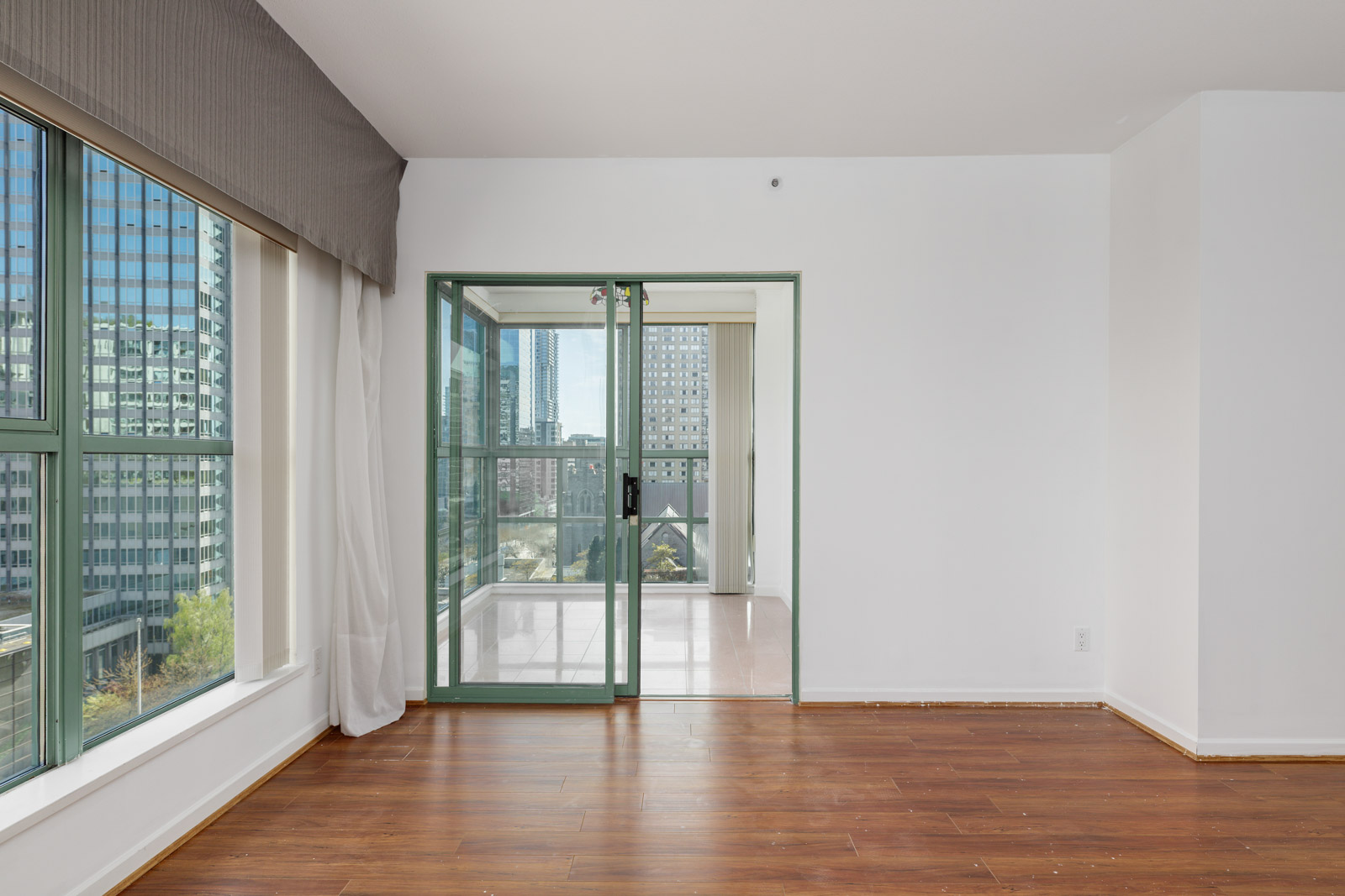 A bright, unfurnished room with large windows, wooden floor, and glass doors leading to an enclosed balcony with a city view.