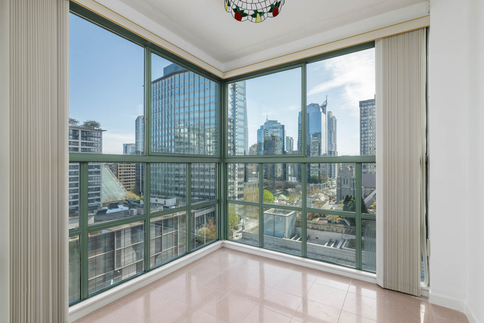 Corner room with large floor-to-ceiling windows, vertical blinds, and a city skyline view with modern high-rise buildings.
