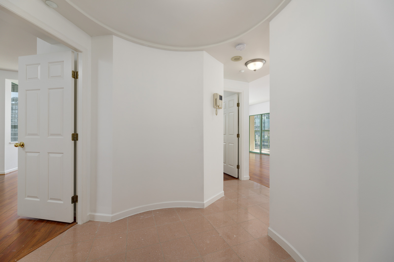 A hallway with light brown tiled floors, white walls, and several open doors leading to rooms with wooden flooring and large windows.