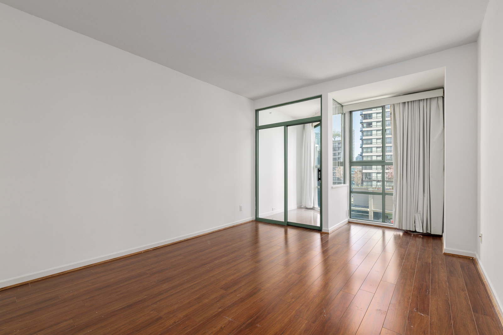 Empty room with white walls, wood flooring, a mirrored closet, and large windows with city views and light-colored curtains.