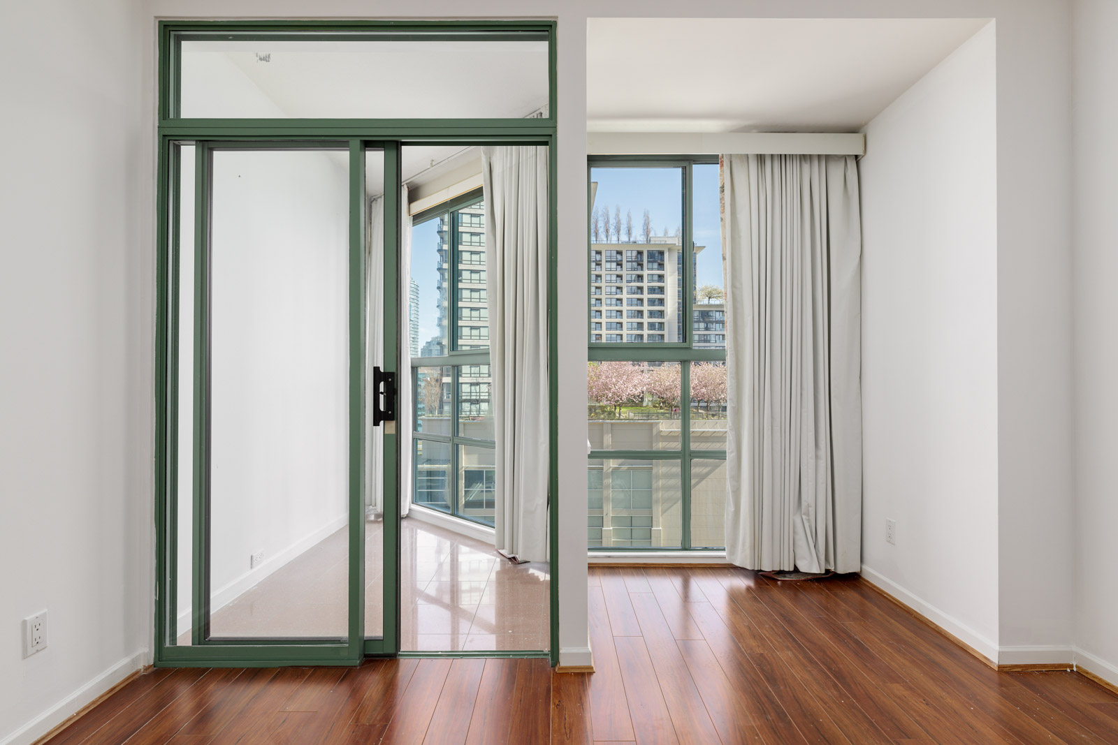 A room with wood floors, white walls, large windows with light curtains, and a glass door leading to a balcony with city buildings visible outside.