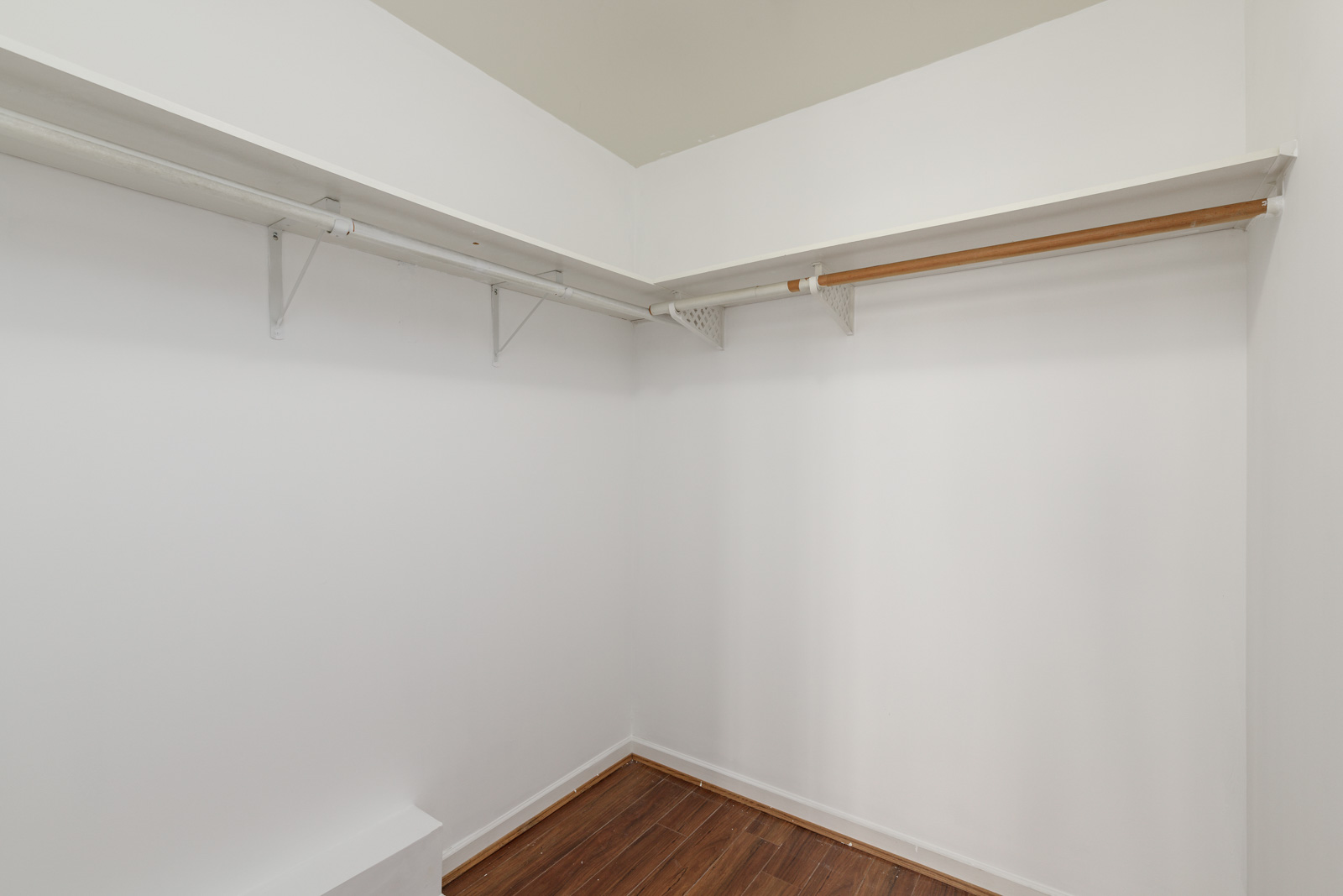 Empty walk-in closet with white walls, wooden floor, and two long shelves with hanging rods along adjacent walls.