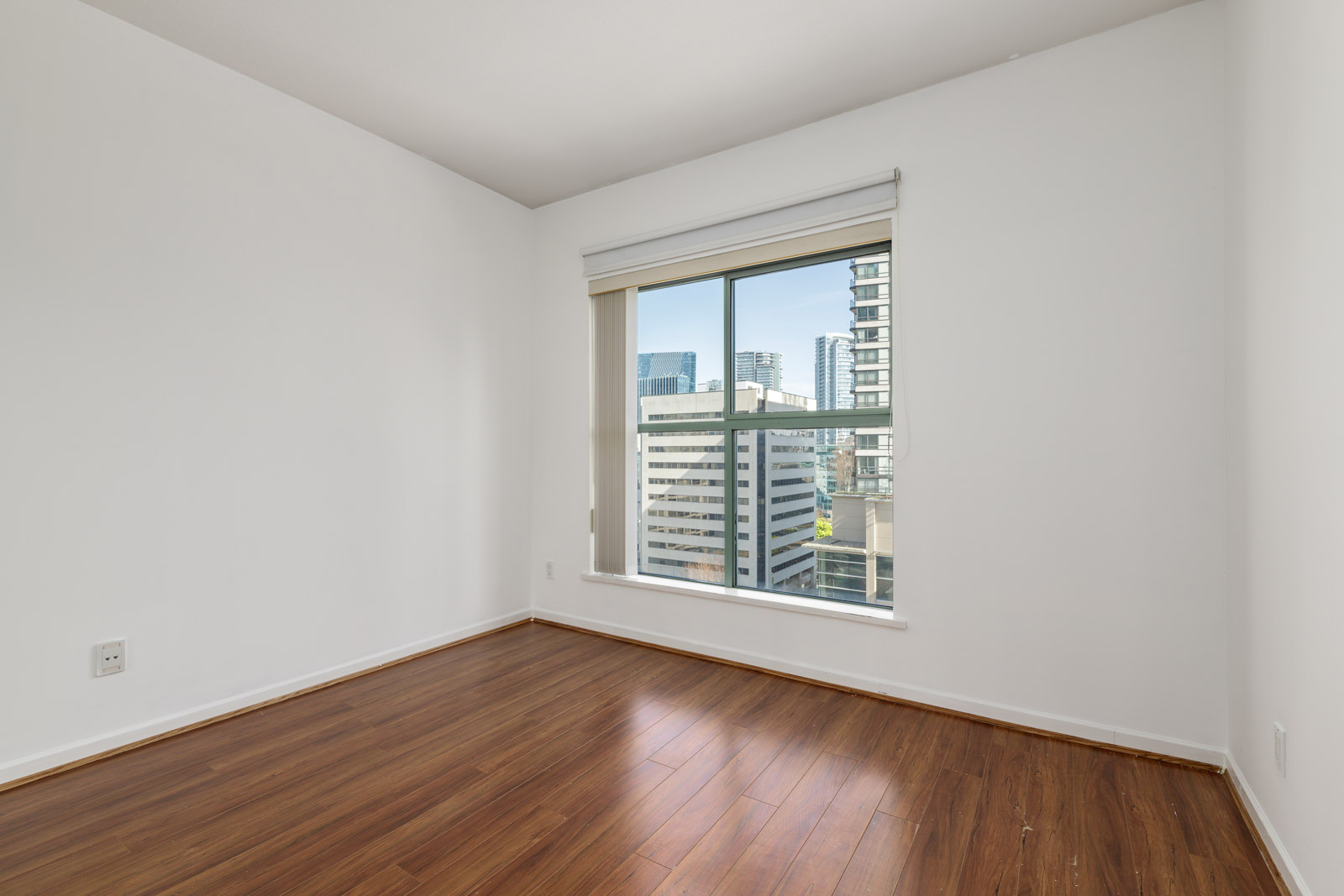 Empty room with white walls, wood flooring, and a large window showing a cityscape with tall buildings outside.