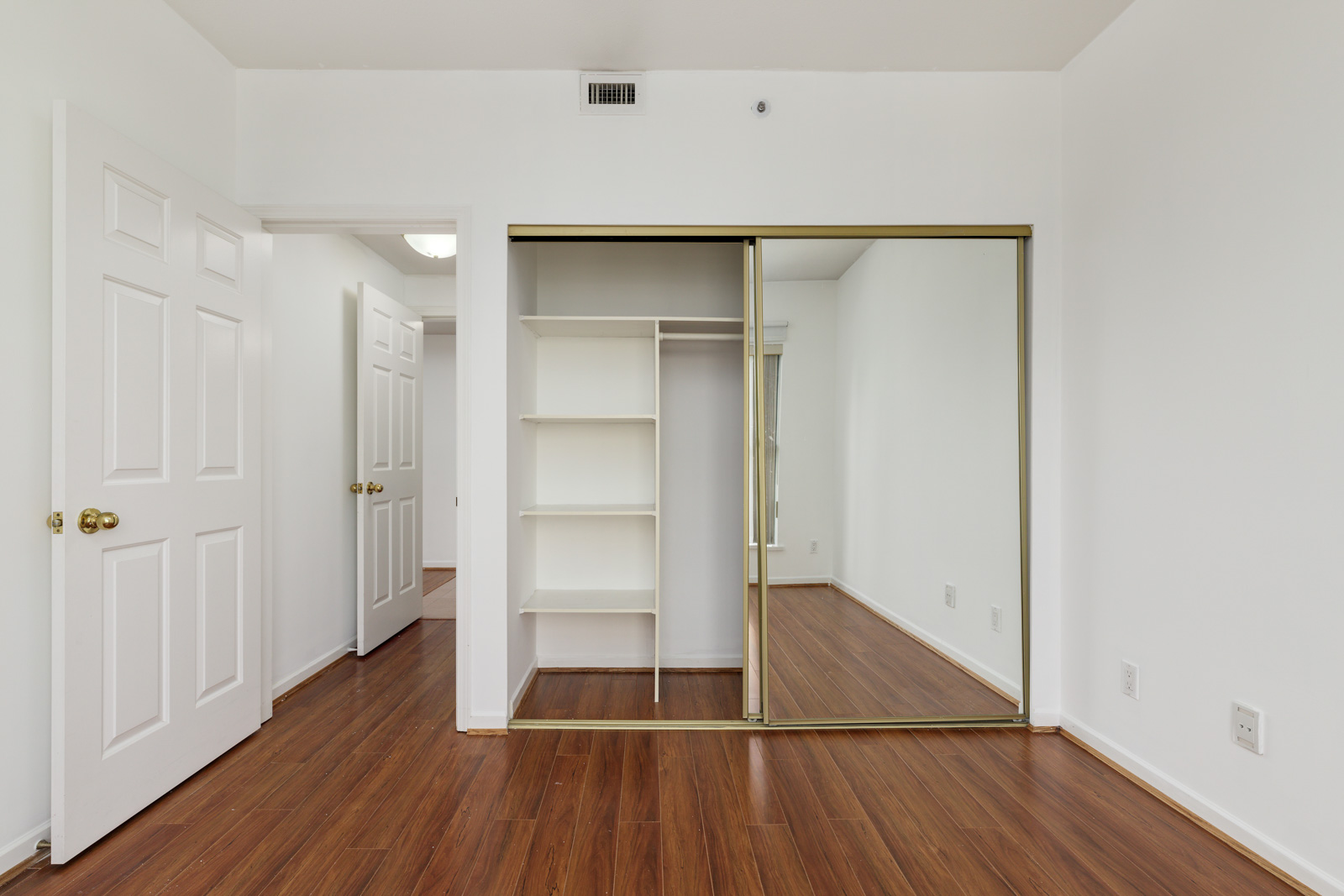 Empty room with white walls, wooden floor, an open closet with shelves, sliding mirrored doors, and two white doors leading to other rooms.