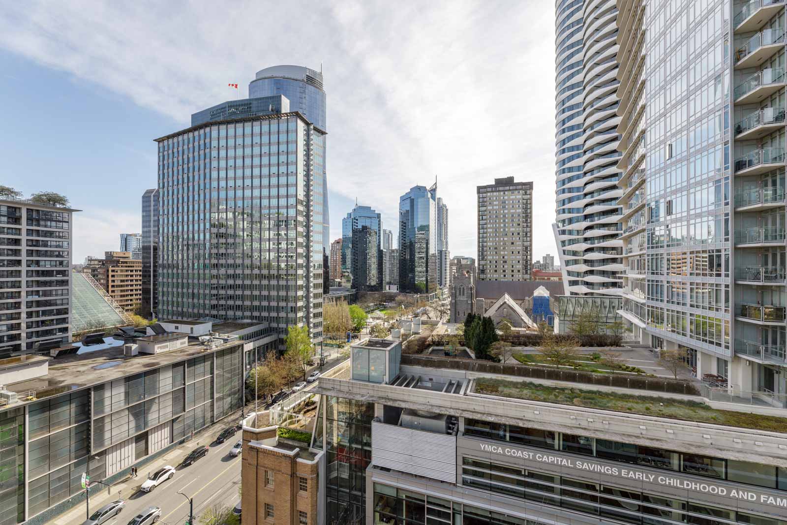Cityscape view featuring modern high-rise buildings, glass facades, and busy streets under a partly cloudy sky, with green rooftop spaces visible in the foreground.