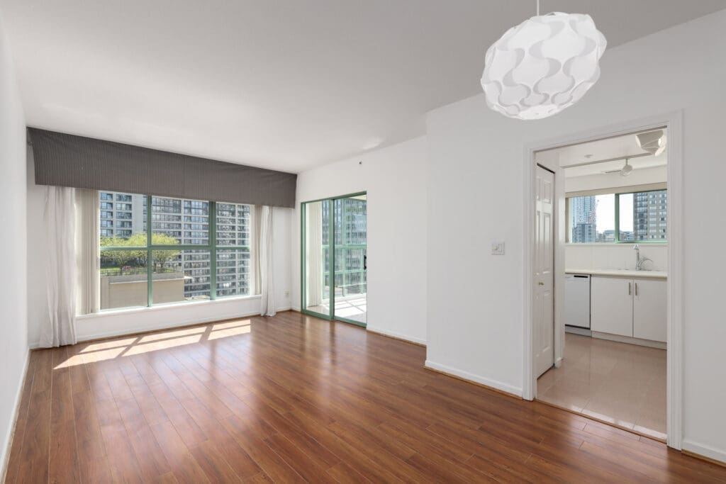 Bright, empty apartment with wood floors, large windows, a sliding glass door to a balcony, and a view into a white kitchen. High-rise buildings visible outside.