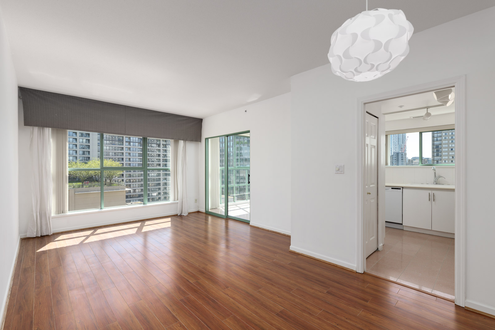 Bright, empty apartment with wood floors, large windows, a sliding glass door to a balcony, and a view into a white kitchen. High-rise buildings visible outside.