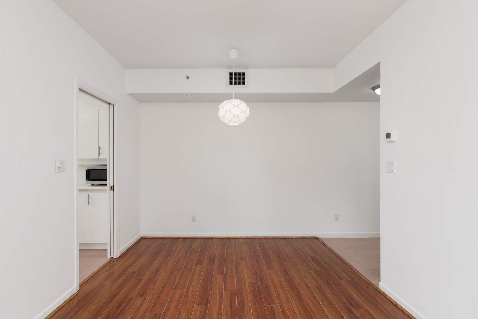 Empty room with white walls, hardwood flooring, a hanging light fixture, and a view into a small white kitchen with a microwave.