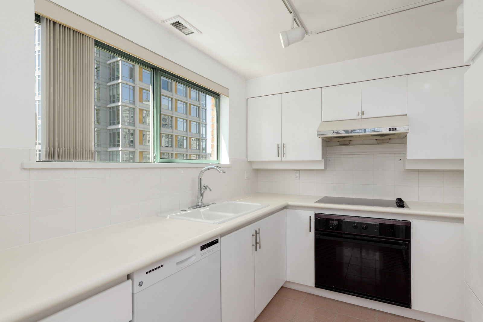 Modern kitchen with white cabinets, a double sink, dishwasher, electric stove, oven, and large window overlooking tall buildings.