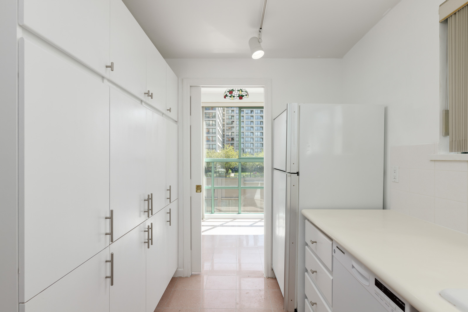 A narrow kitchen with white cabinets, a white refrigerator, and a view of a balcony and tall buildings through a glass door at the far end.