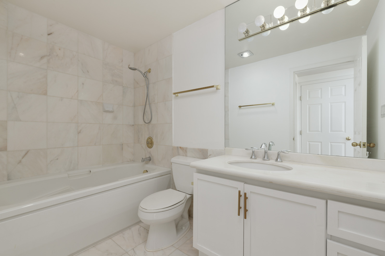 Modern bathroom with white marble tiles, bathtub with shower, toilet, white vanity with sink, large mirror, and overhead light fixture.