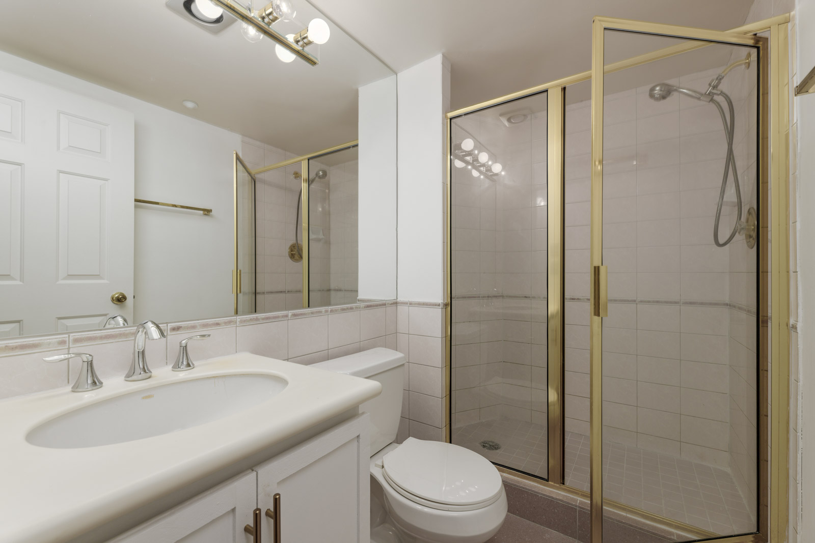 Bathroom with white sink, toilet, and a corner glass shower with gold trim. A large mirror and overhead lights are visible above the vanity.