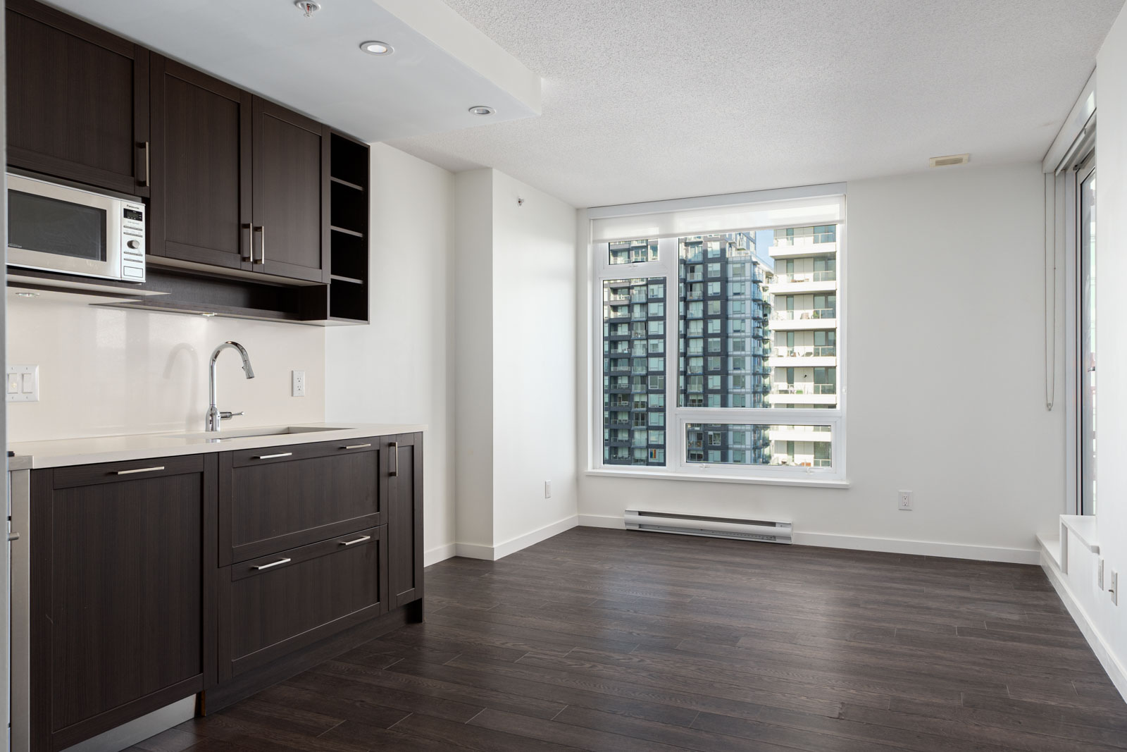 Modern apartment with dark wood kitchen cabinets, stainless steel appliances, and large windows overlooking city buildings. White walls and dark wood floors throughout.
