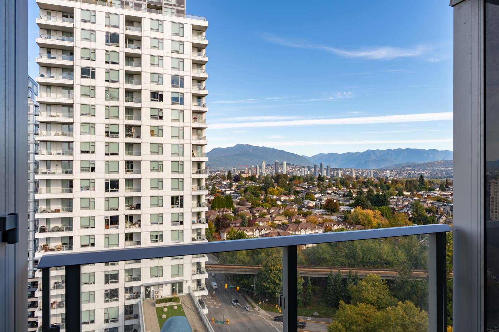 View from a balcony overlooking a cityscape with a tall apartment building, residential houses, trees, and mountains in the background under a clear blue sky.
