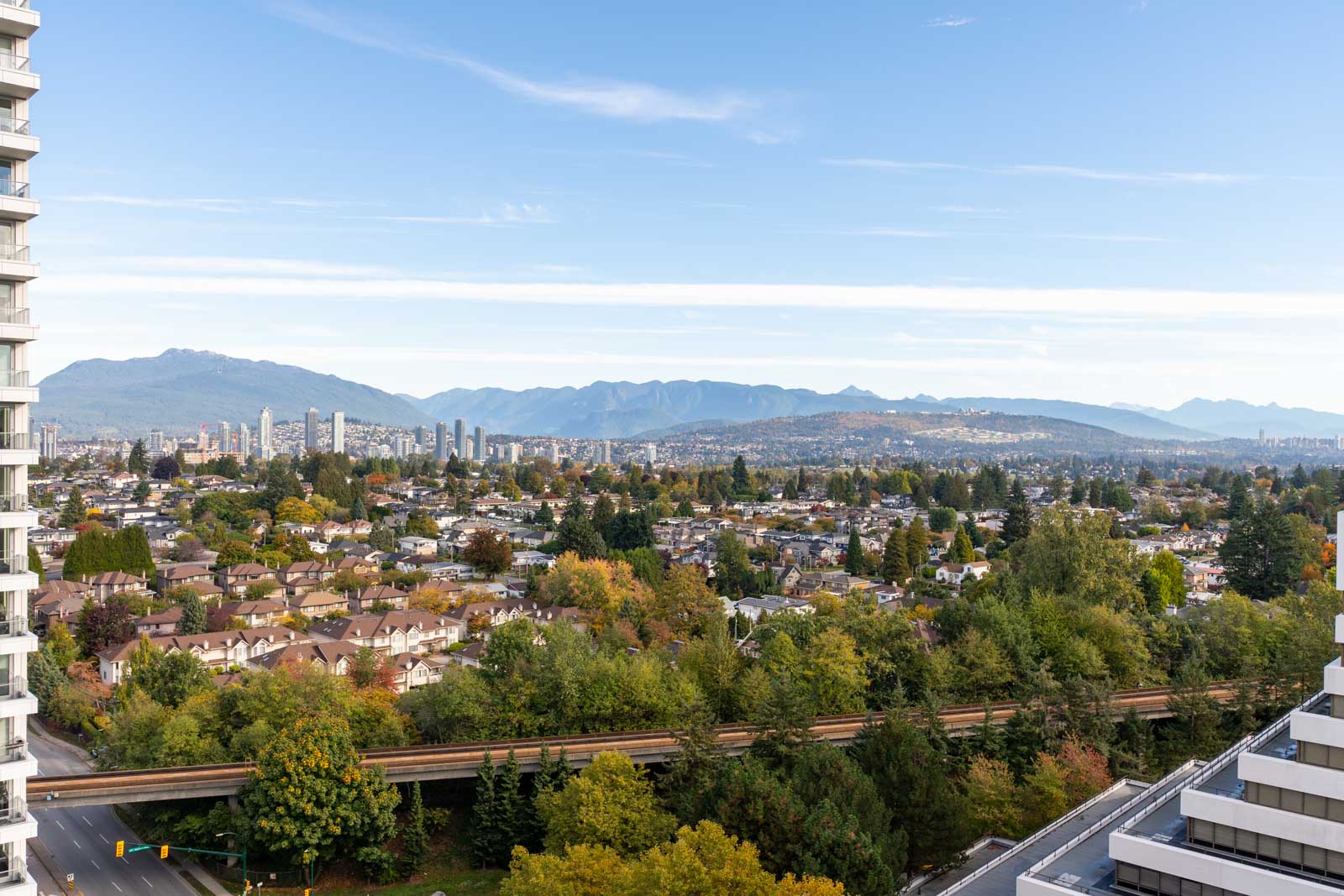 View from a high-rise building overlooking suburban houses, a light rail track, and distant city skyline with mountains in the background under a clear sky.