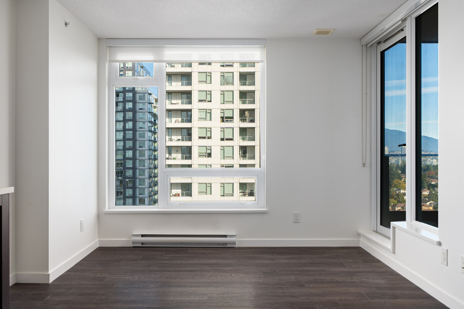 Empty modern room with white walls, dark wood flooring, large windows showing a city apartment building and partial mountain view.