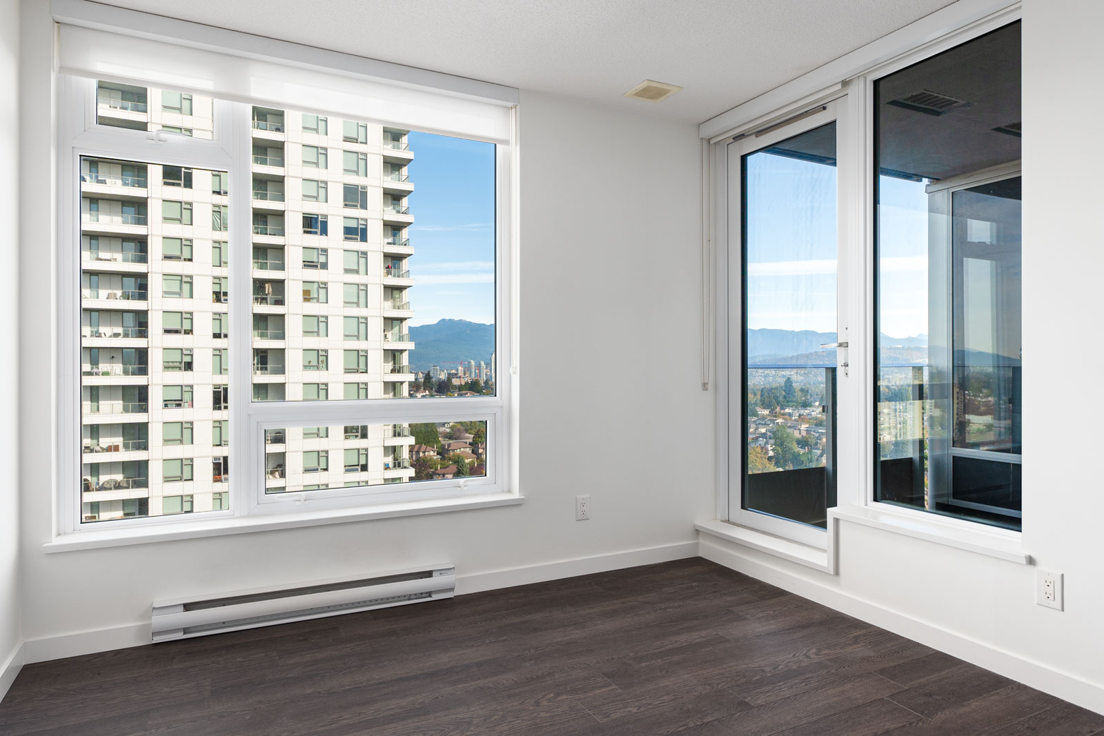 Empty modern apartment room with large windows, dark wood flooring, and a glass door leading to a balcony with views of nearby buildings and distant mountains.