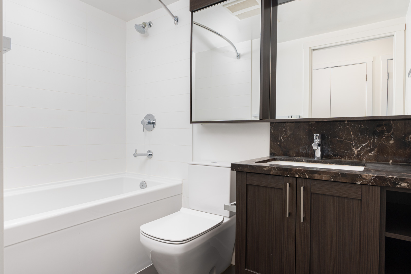 Modern bathroom with a bathtub, toilet, dark wood vanity with sink, marble countertop, and mirrored cabinet above.