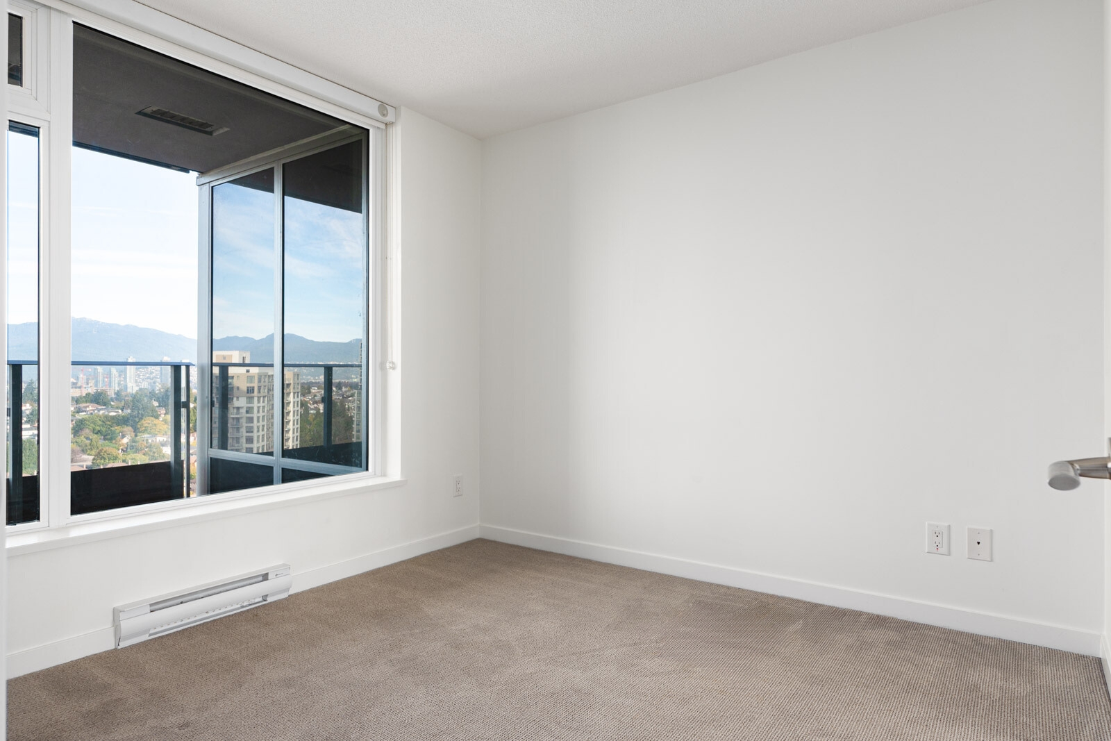 Empty room with beige carpet, white walls, large window, and glass door leading to a balcony with a city and mountain view.
