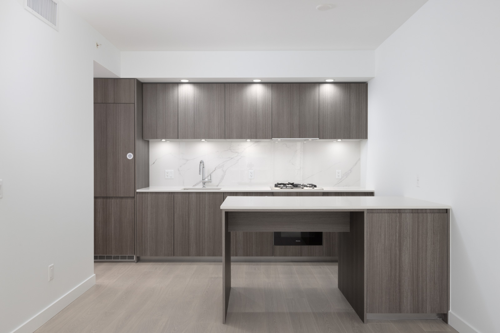 Modern kitchen with gray wood cabinets, built-in stove, marble backsplash, under-cabinet lighting, and a peninsula counter. The space features light wood flooring and white walls.