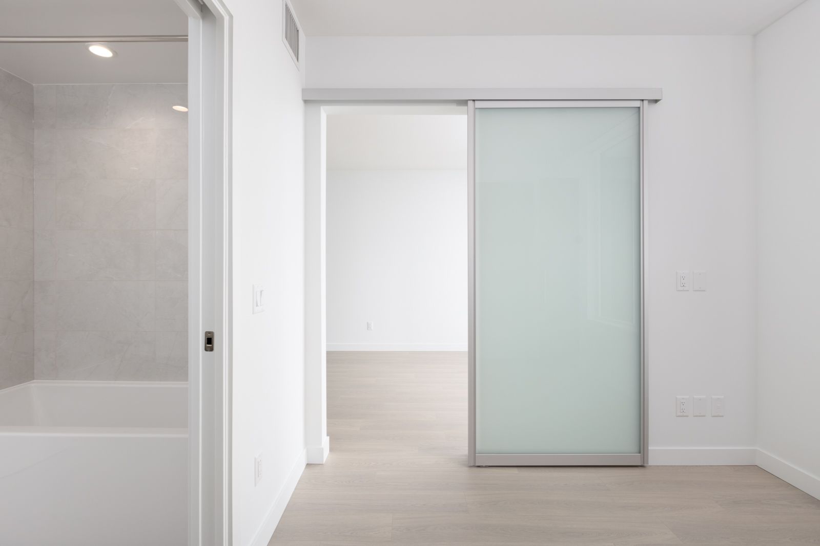 Minimalist interior with a frosted glass sliding door, light wood floor, white walls, and a bathroom with gray tiles and a bathtub visible on the left.