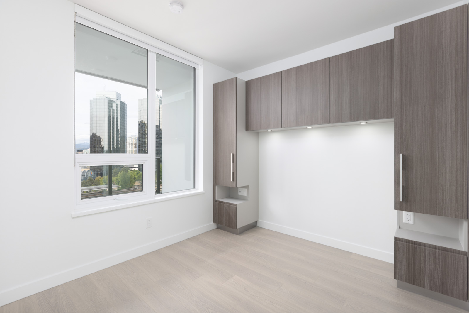 A modern, unfurnished room with light wood flooring, built-in wood cabinets, large window, and a city view.