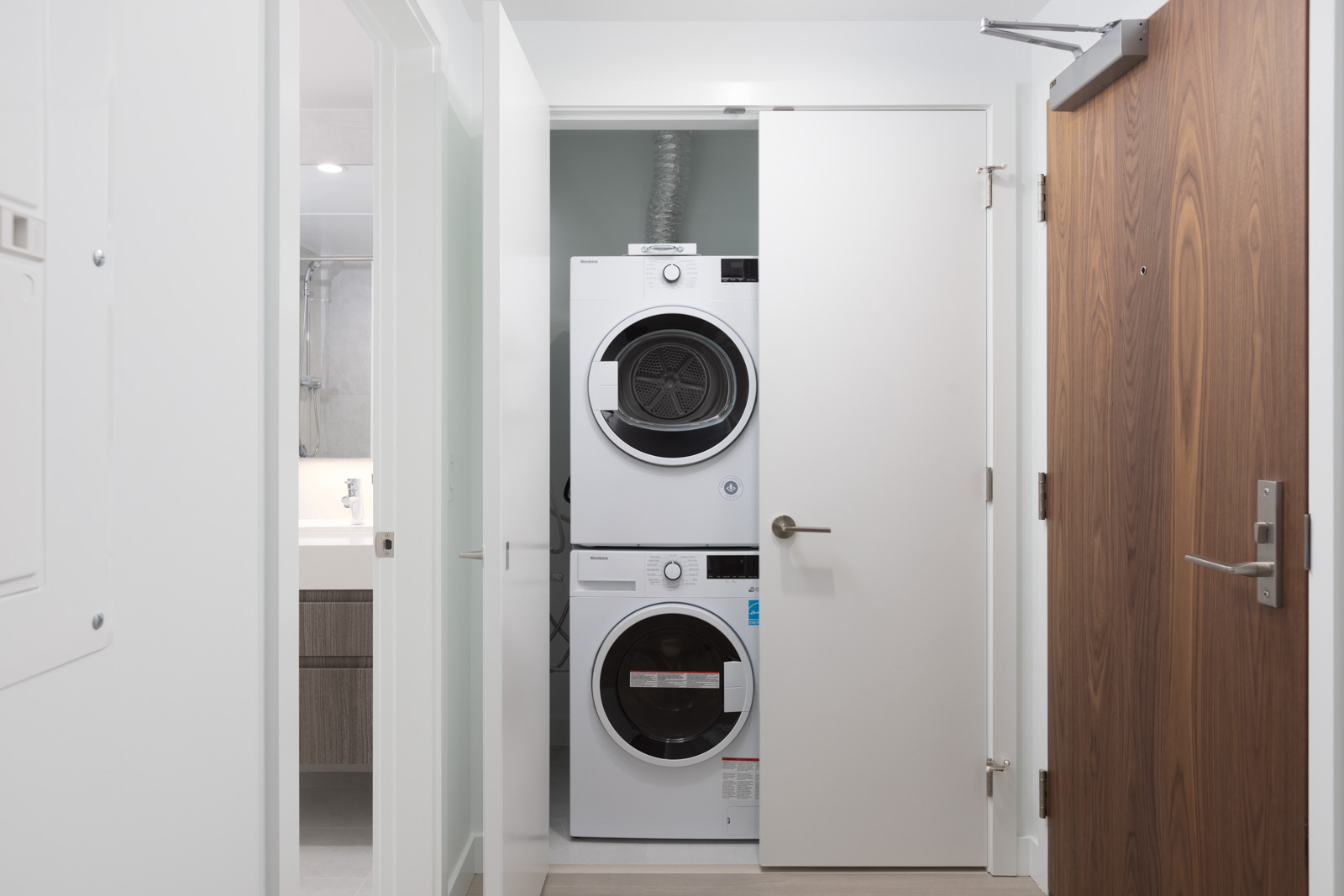 A stacked washer and dryer set is installed in a closet space with a partially open white door in a modern apartment hallway.
