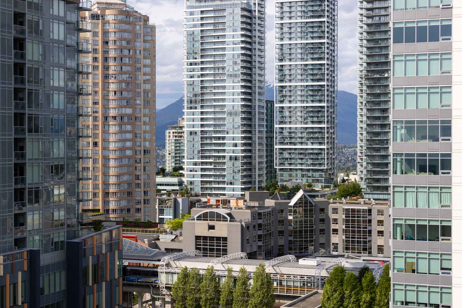 Tall modern glass and concrete high-rise buildings grouped together in an urban cityscape, with trees and mountains visible in the background.