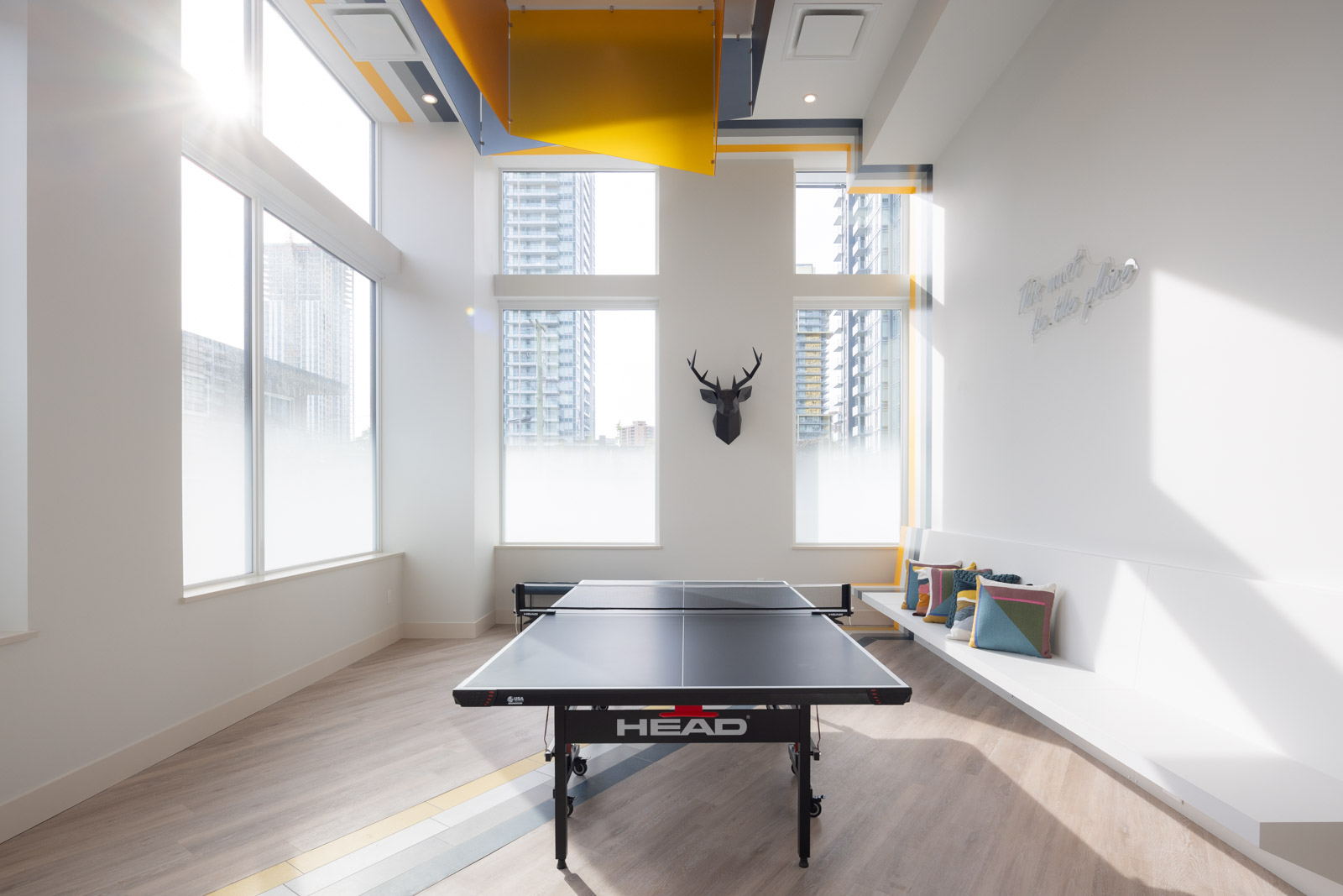A modern, brightly lit room with large windows, a black HEAD ping pong table in the center, a wall-mounted deer head, and built-in bench seating with cushions.