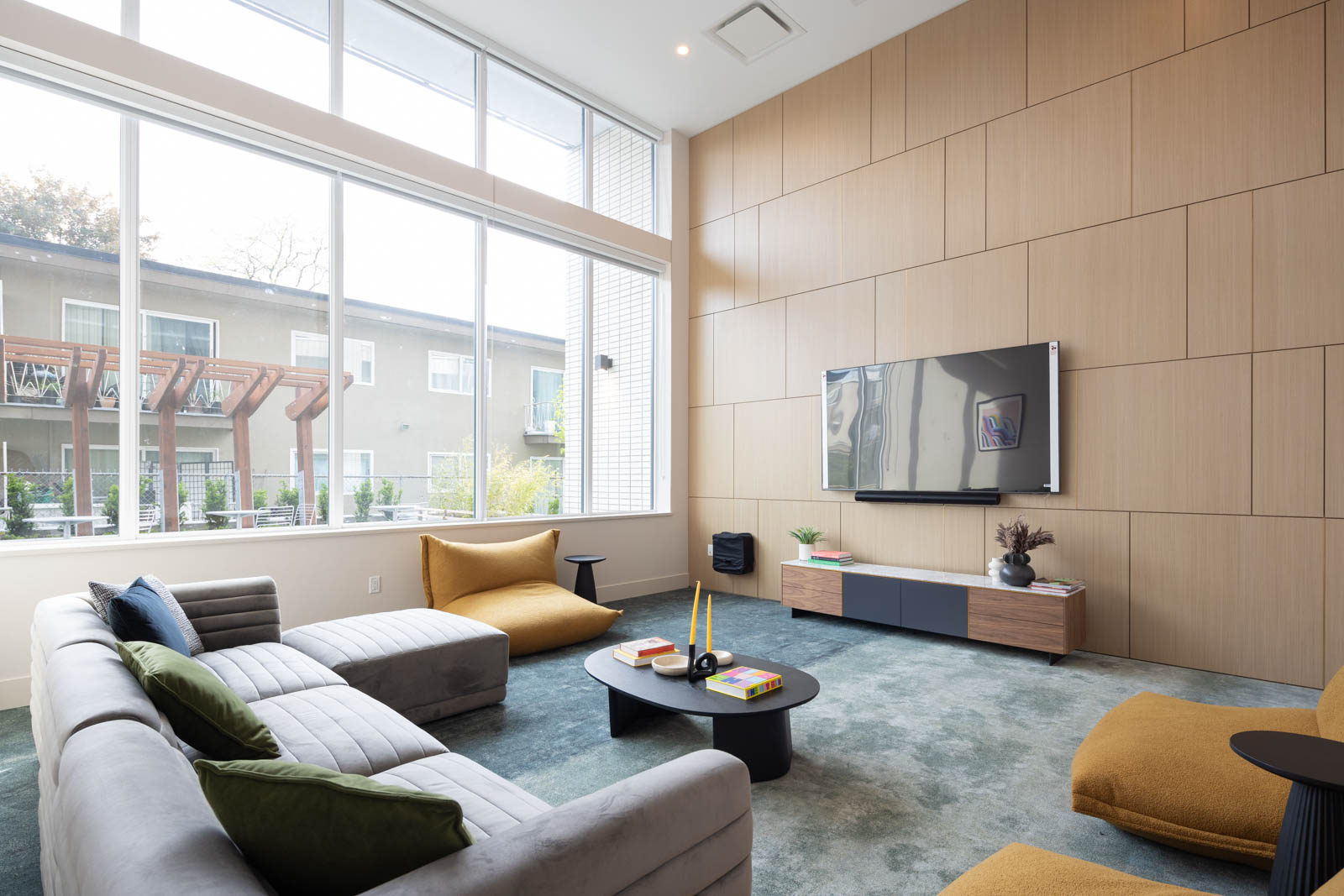 Modern living room with large windows, gray sectional sofa, two mustard chairs, coffee table with books, and wall-mounted TV on wood-paneled wall.