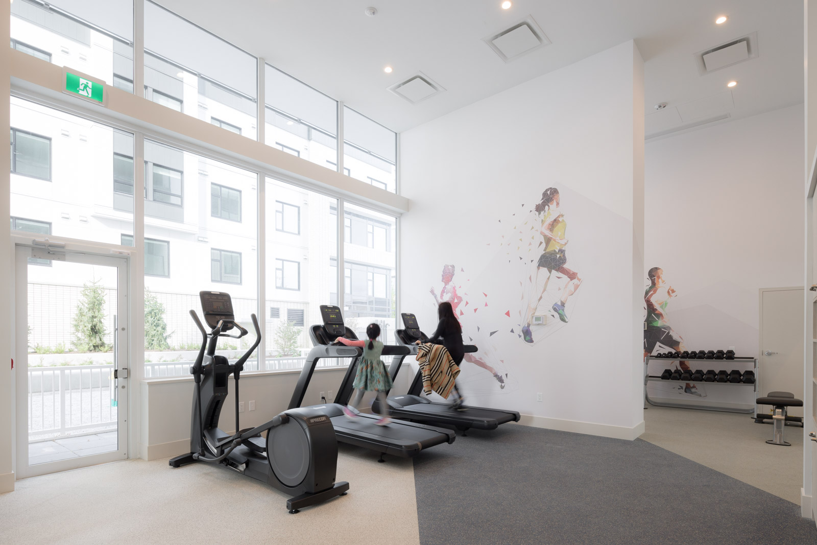 Two people use treadmills in a bright gym with large windows, fitness equipment, and murals of runners on the walls.