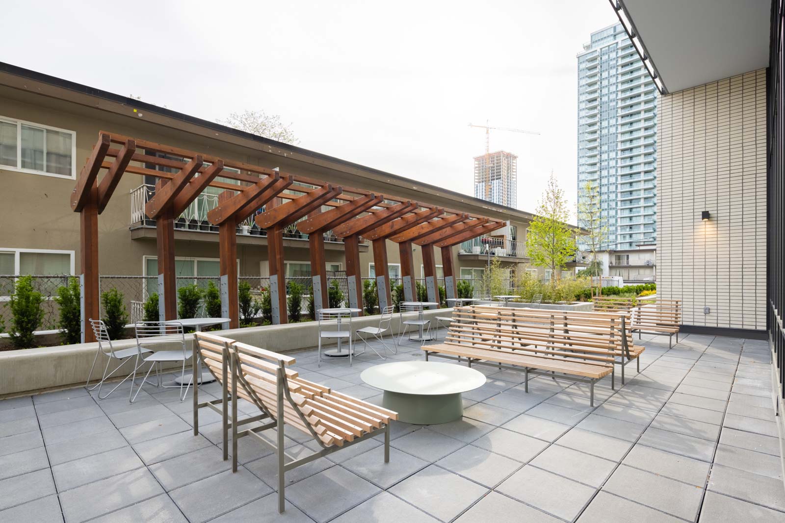 Outdoor patio area with wooden benches, metal chairs, a round table, pergola, and surrounding apartment buildings in the background.