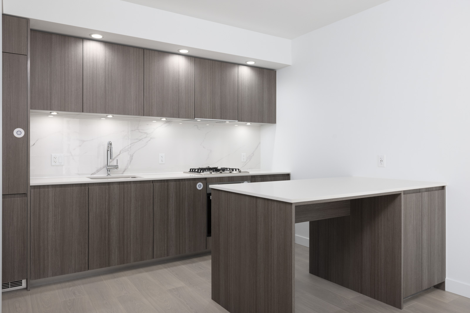 Modern kitchen with wood cabinets, marble backsplash, built-in gas stove, under-cabinet lighting, and a white island counter on a light wood floor.