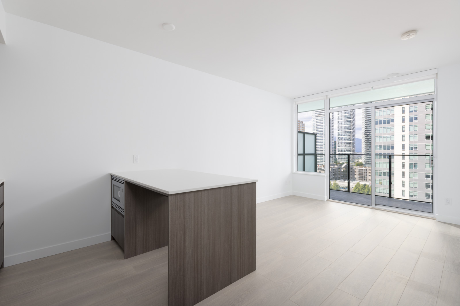 Modern, unfurnished apartment interior with light wood floors, a kitchen island, and large windows leading to a balcony with a city view.