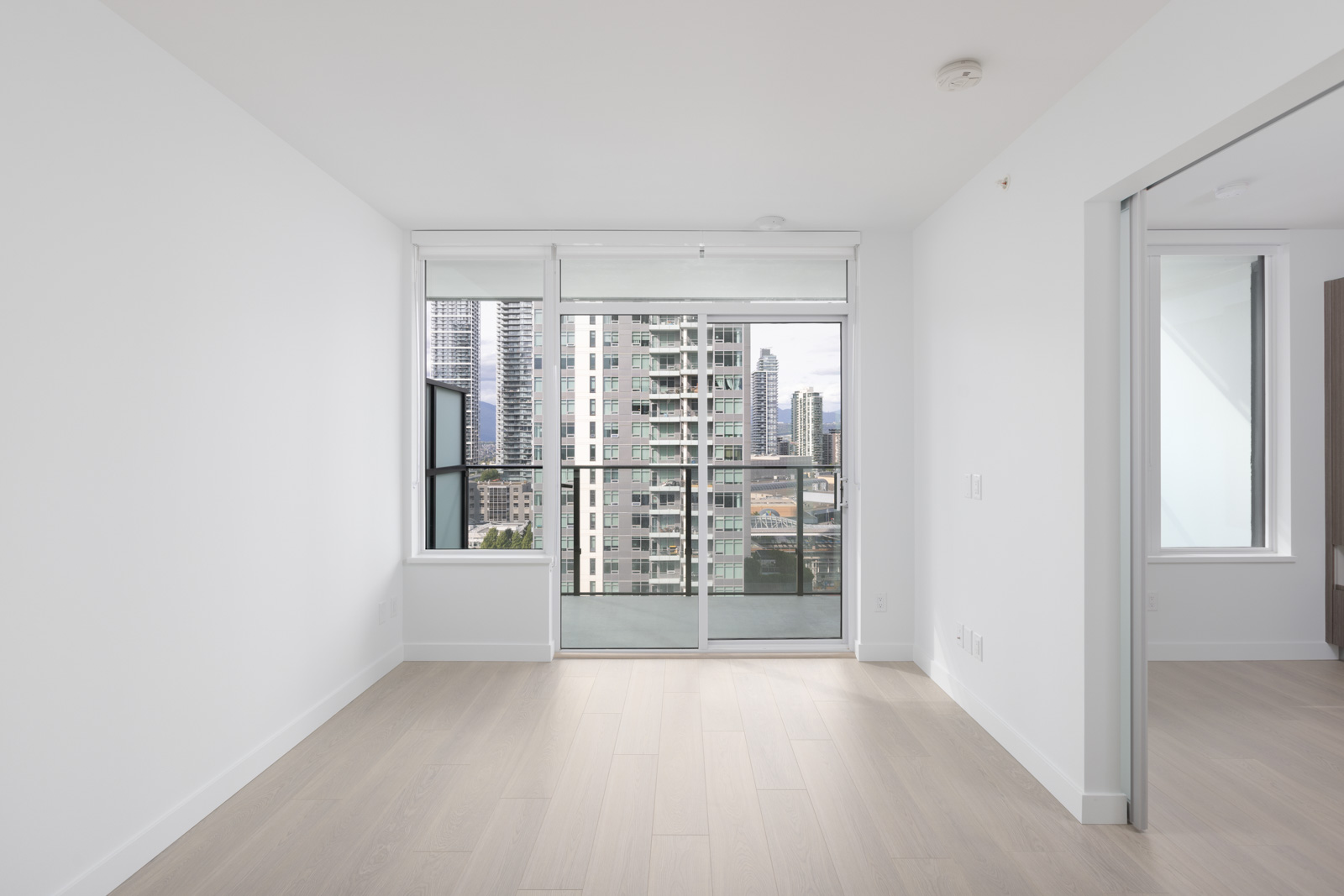 Empty modern apartment room with light wood floors, white walls, large window, and glass door opening to a balcony with city buildings visible outside.