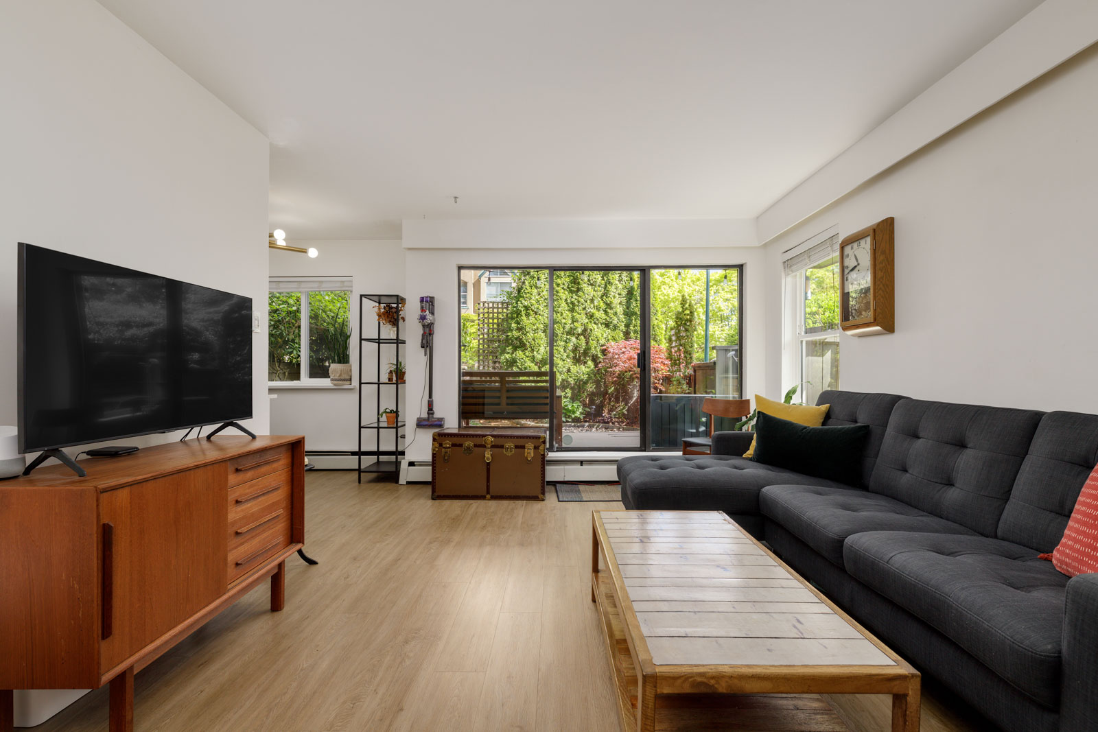 Modern living room with a black sofa, wooden coffee table, TV on a wooden stand, and large glass doors opening to a patio with greenery outside.