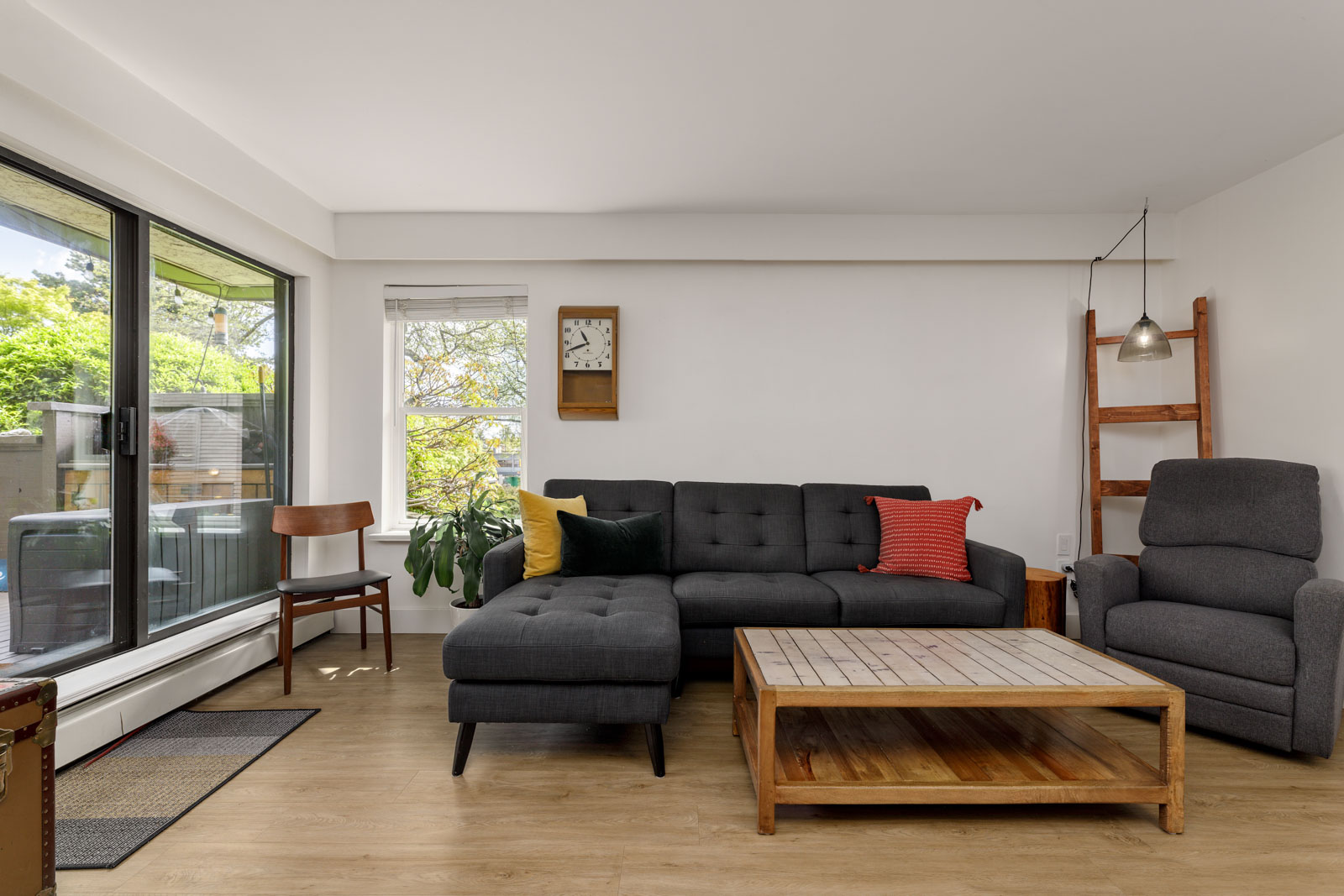 Modern living room with a dark grey sectional sofa, wooden coffee table, armchair, chair, clock, and large window showing greenery outside.